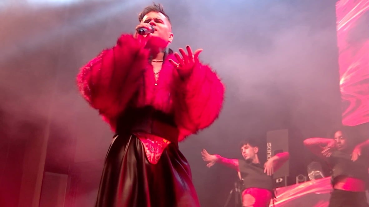 Lui da Motta in red fur, arms raised, mid-song under pink light.
